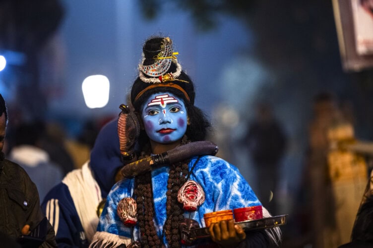 A person dressed as a deity with blue face paint and traditional attire holds a tray, embodying the vibrant spiritual essence of Varanasi, India.