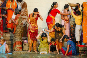 A group of people, including women and children, gather at a riverbank in Varanasi. One woman pours water over her head while others stand and interact. They're dressed in colorful traditional attire.