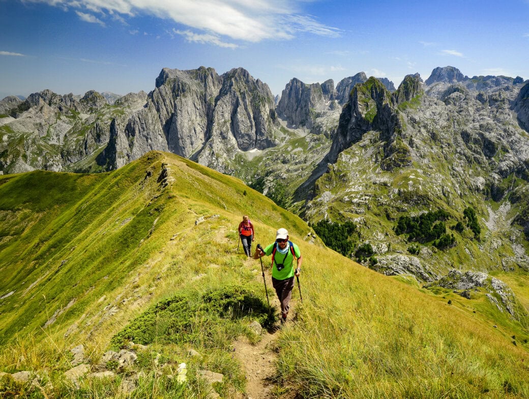 Two hikers with backpacks and walking poles ascend a grassy mountain ridge against a backdrop of rugged peaks under a clear blue sky.