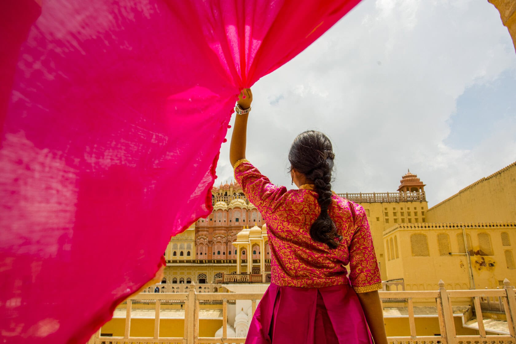 In a pink traditional attire, a person holds vibrant fabric, gazing at a historic building under Rajasthan's cloudy sky.