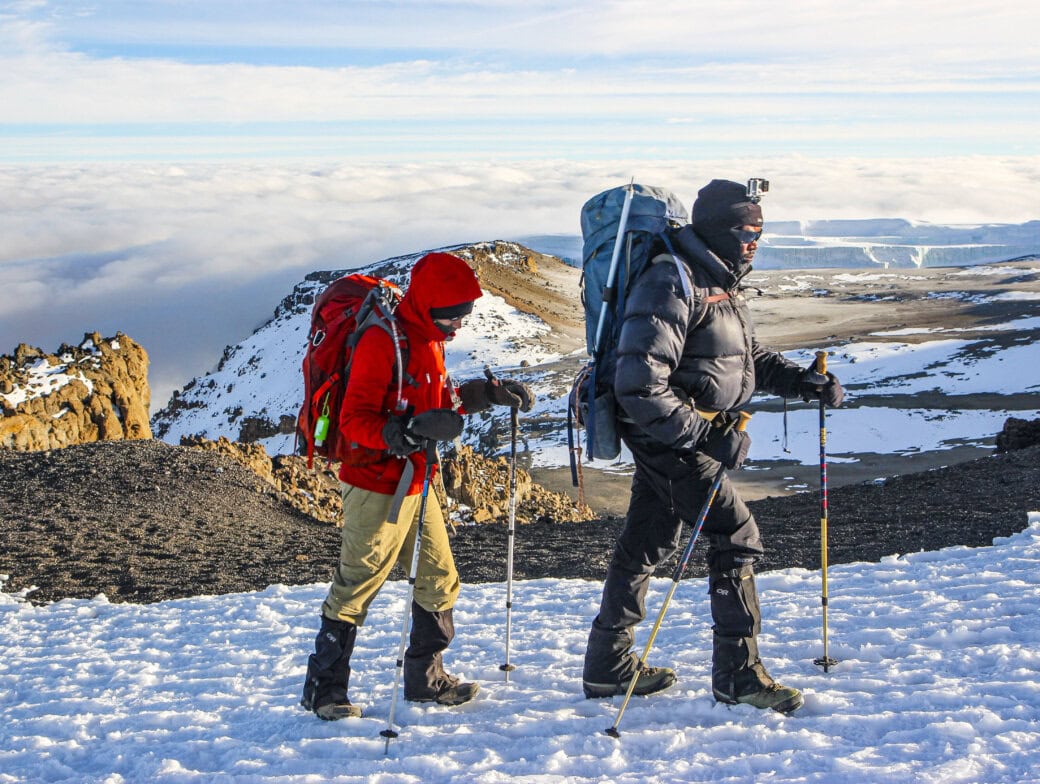 Two hikers in cold-weather gear walking on a snow-covered mountain path against a backdrop of clouds and rocky terrain.