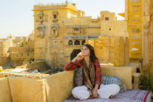 A person sitting cross-legged on a cushioned terrace in Rajasthan, looking up thoughtfully. Historic sandstone buildings rise majestically in the background under a clear sky, making this moment feel like it belongs in a beginner's guide to India.