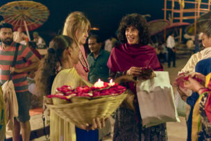 Amidst the bustling night market of Varanasi, people gathered under a starlit sky, with a woman gracefully holding a basket of flowers and candles in the foreground.