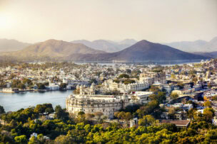 Aerial view of Udaipur, Rajasthan, showcasing historic architecture, lush greenery, a large palace, and distant hills under a hazy sky—a perfect introduction for any beginner's guide to India's beauty.