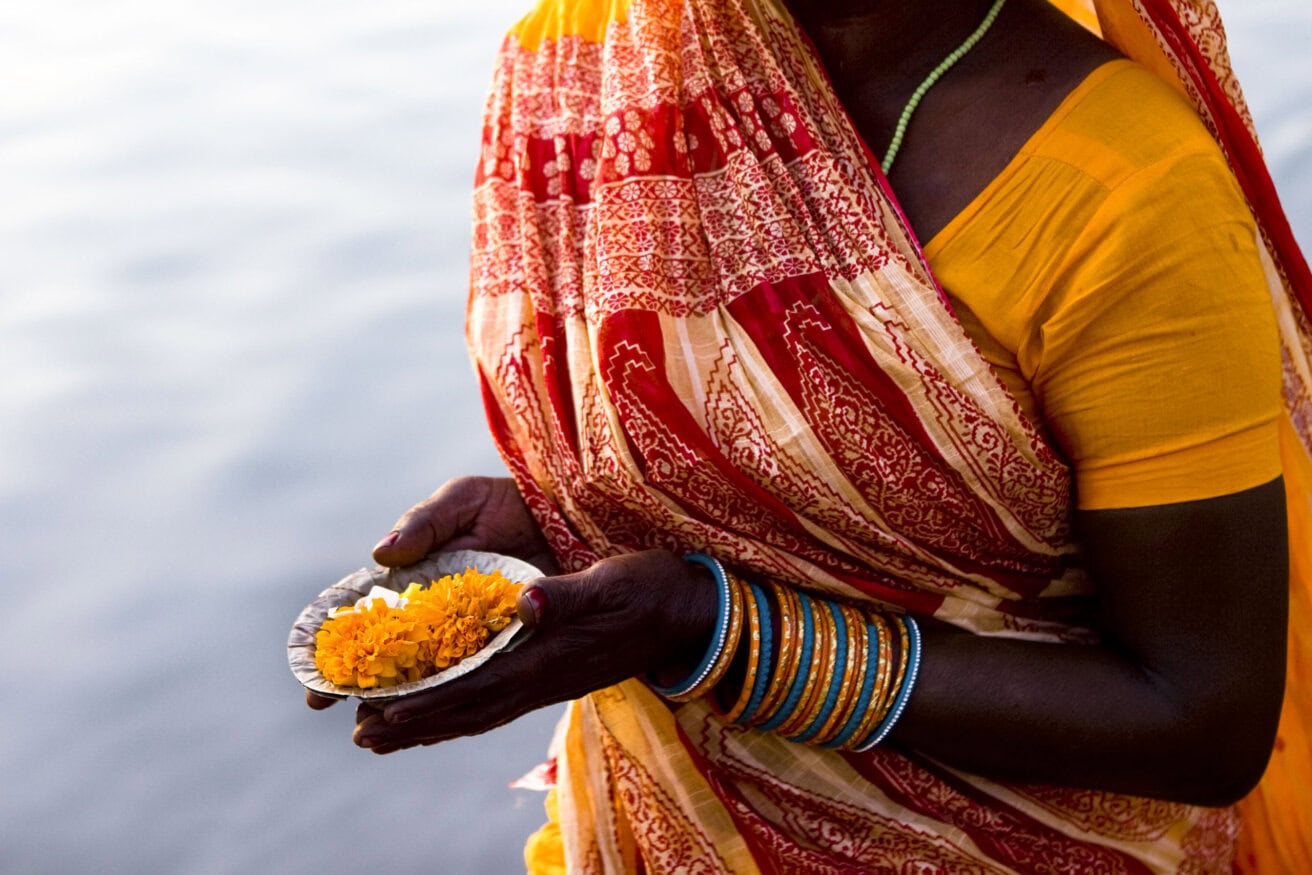 A person in a colorful sari holds a small dish with marigold flowers, standing near the sacred waters of Varanasi.