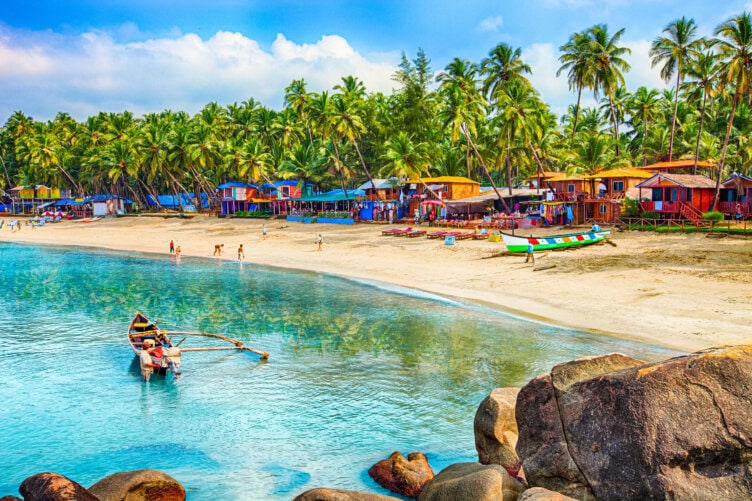 A colorful beach scene reminiscent of India's coastal charm, with palm trees, huts, and boats along a sandy shoreline. People wade in the clear water, exploring the rocks in the foreground—one of the must-do experiences for anyone seeking tranquility by the sea.