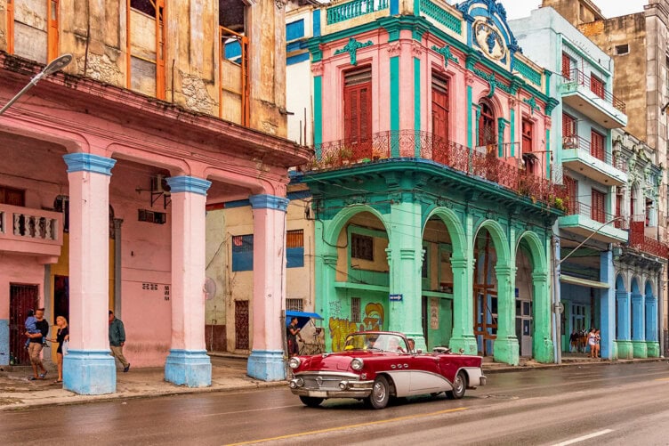 A classic car glides through the vibrant streets of Cuba, past colorful, weathered buildings with arches.