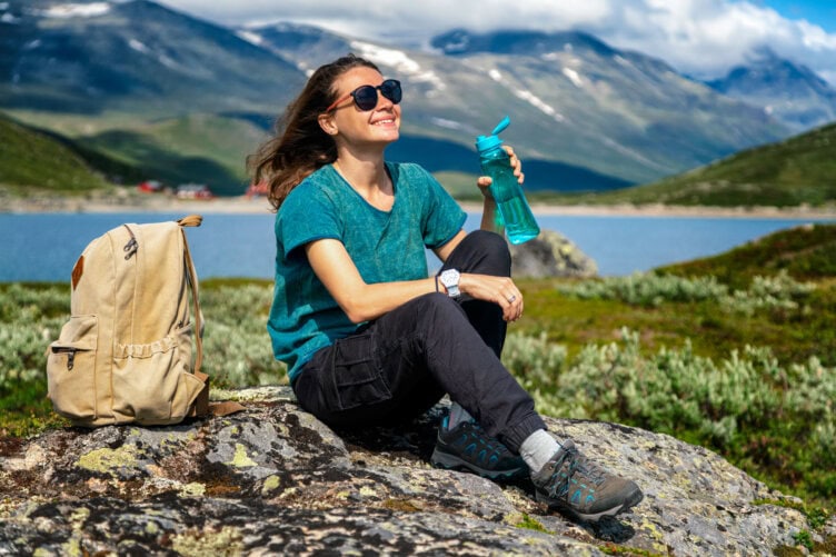 A person sits on a rock by the lake, clad in casual hiking attire and sunglasses. Their trusty hiking boots rest nearby next to a beige backpack. They sip from a water bottle, with majestic mountains and clouds providing the perfect backdrop for this serene moment.
