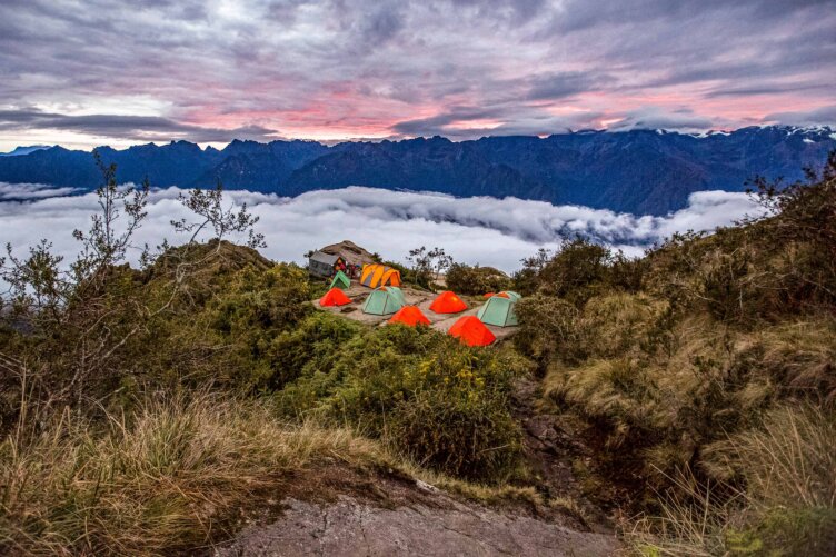 Campsite with several tents perched on a mountain ridge, offering breathtaking views of a cloud-filled valley. Rugged peaks rise dramatically against the colorful sunset sky, reminiscent of scenes from the iconic Inca Trail's 5-day journey.