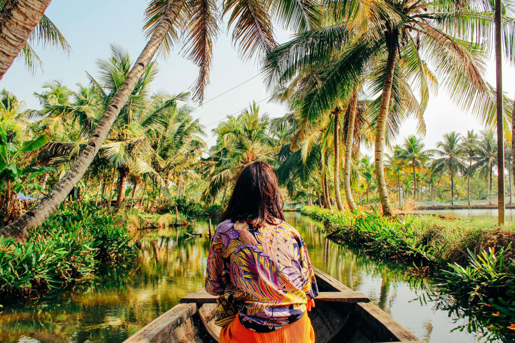 Sporting a colorful shirt, the traveler with long hair relaxes in a boat drifting along a tropical waterway lined with palm trees—a quintessential must-do for anyone exploring the serene beauty of India.