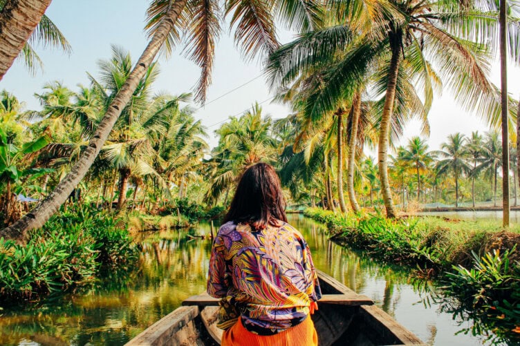 Sporting a colorful shirt, the traveler with long hair relaxes in a boat drifting along a tropical waterway lined with palm trees—a quintessential must-do for anyone exploring the serene beauty of India.