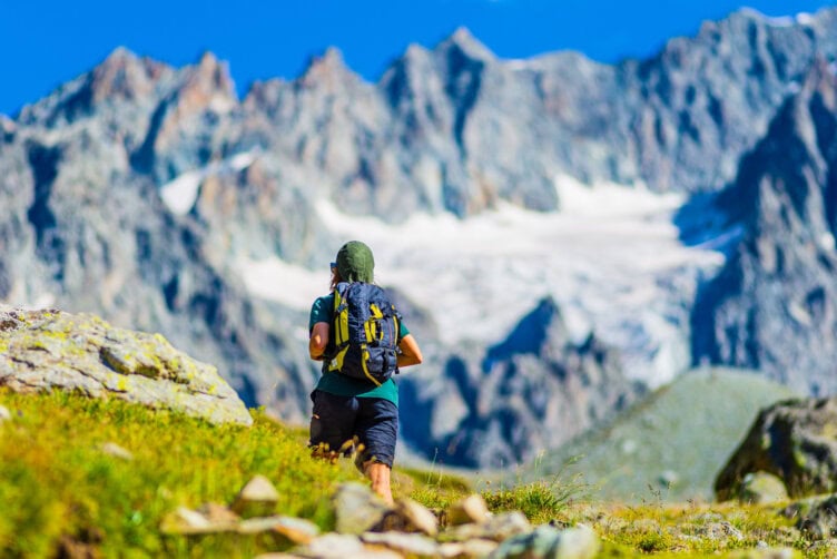 A person wearing a travel backpack hikes along a grassy trail, set against the majestic backdrop of the rocky Alps under a clear blue sky.