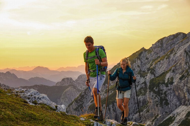 Two hikers with poles ascend a mountain trail in the Alps at sunset, surrounded by rocky peaks and a vibrant sky, their pack filled with travel essentials.