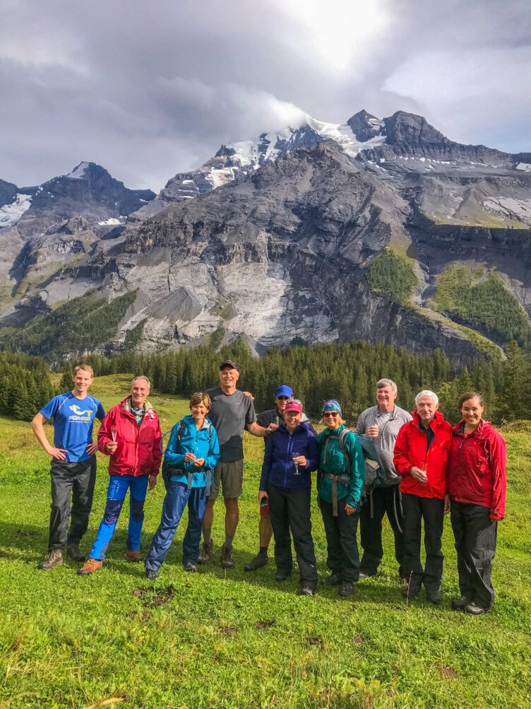 A group of nine people in outdoor gear stands on a grassy area with a backdrop of rugged, snow-capped Alps and forest, having carefully prepared their travel essentials for the adventure ahead.