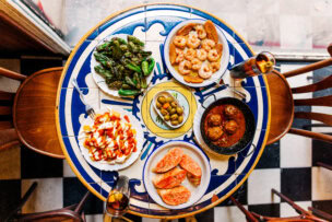A table brimming with Madrid's top tapas: Padrón peppers, shrimp, olives, meatballs, tomato bread, and patatas bravas. It's one of the top things to do in Madrid, with each dish perfectly paired alongside two refreshing glasses of iced drinks.