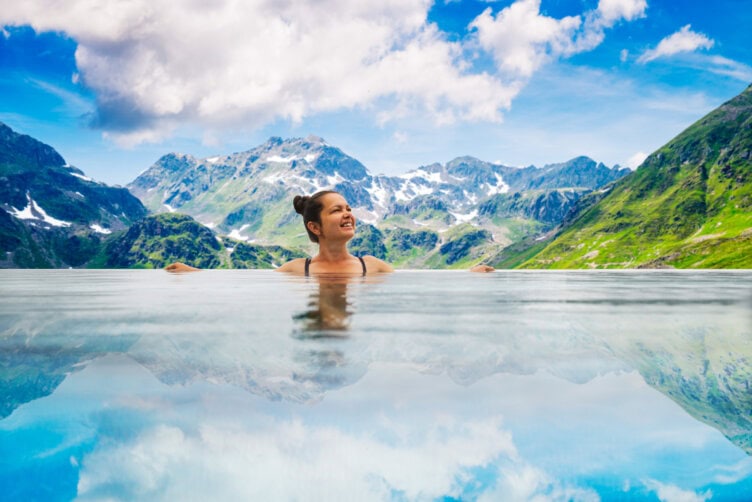 Woman smiling in an infinity pool with a breathtaking view of the Alps, their peaks rising majestically under a partly cloudy sky. It's the perfect travel moment in a serene setting.