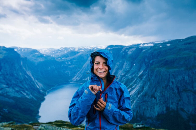 A person smiles, wrapped snugly in a blue jacket with a hood, standing amidst the breathtaking Alps. Overlooking a serene lake under a cloudy sky, it's the perfect travel moment packed with nature's beauty.