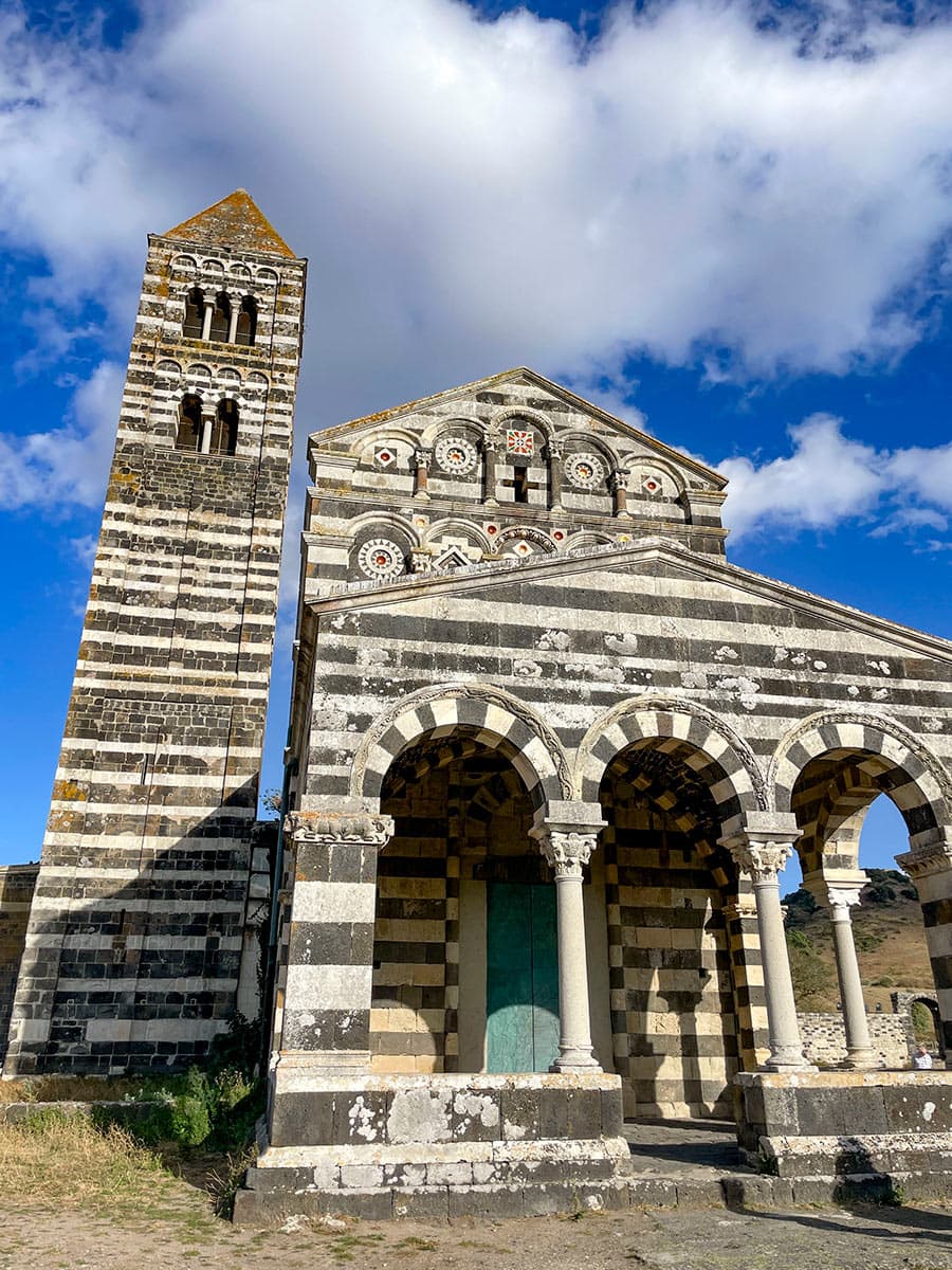 Striped medieval church with an arched facade and tall bell tower stands majestically against a blue sky with clouds, reminiscent of the serene landscapes found while hiking in Sardinia.