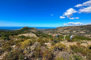 Hikers explore a rocky hillside under a clear blue sky, with the distant mountains of Corsica and the shimmering sea in the background.