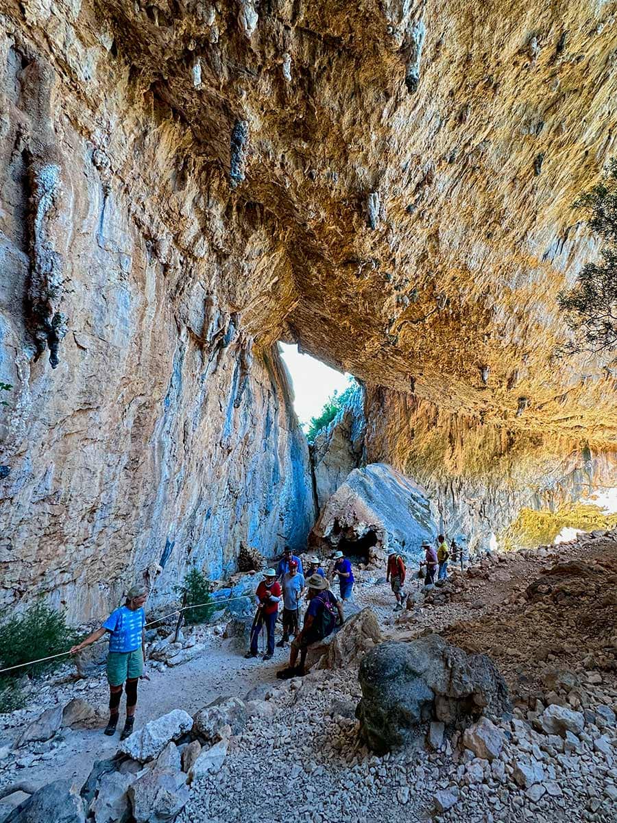 People exploring a large, rocky cave with a high ceiling and natural light streaming through an opening, reminiscent of the stunning landscapes found in Sardinia. Some individuals are standing while others walk along a path, enjoying the serene atmosphere.