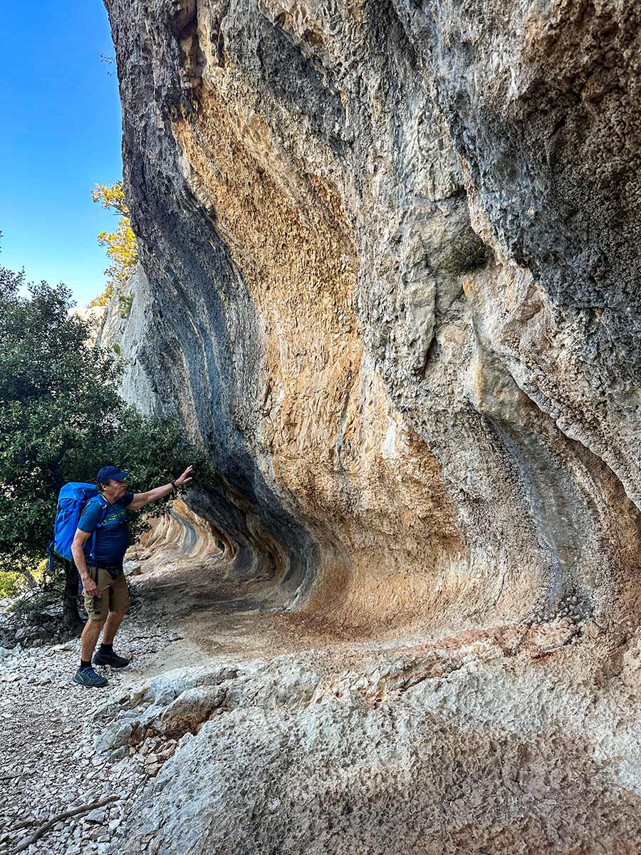 A person with a blue backpack points at a curved rock formation on the hiking trail, surrounded by trees and a clear Sardinian sky.
