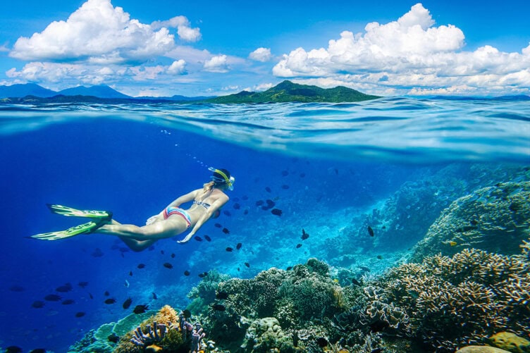 In the clear blue waters of Cenderawasih Bay, a snorkeler glides effortlessly among vibrant coral reefs and colorful fish, with distant islands silhouetted under a partly cloudy July sky.