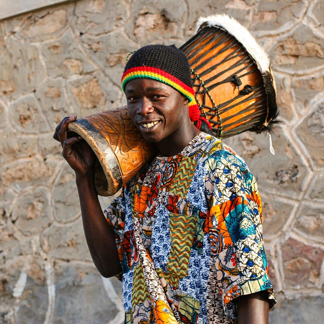 A man in a colorful shirt and hat smiles, holding a decorated drum on his shoulder, inviting you to explore the vibrant rhythms of Cape Verde against a stone wall background.