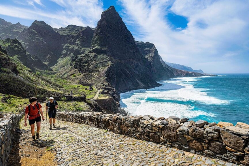 Two people explore a stone path along the mountainous Cape Verde coastline, with ocean waves crashing below under a partly cloudy sky.