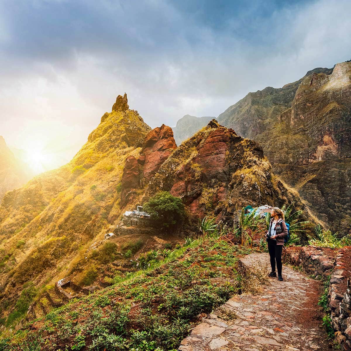 A person walks along a stone path in Cape Verde, surrounded by lush green vegetation and towering, rugged mountains. Under a cloudy sky with sunlight breaking through, the scene is an irresistible invitation to explore the island's natural beauty.