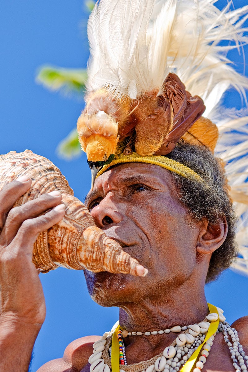 Against the July sky, a person in a feathered headdress blows into a large conch shell, echoing the vibrant sounds of Cenderawasih Bay.