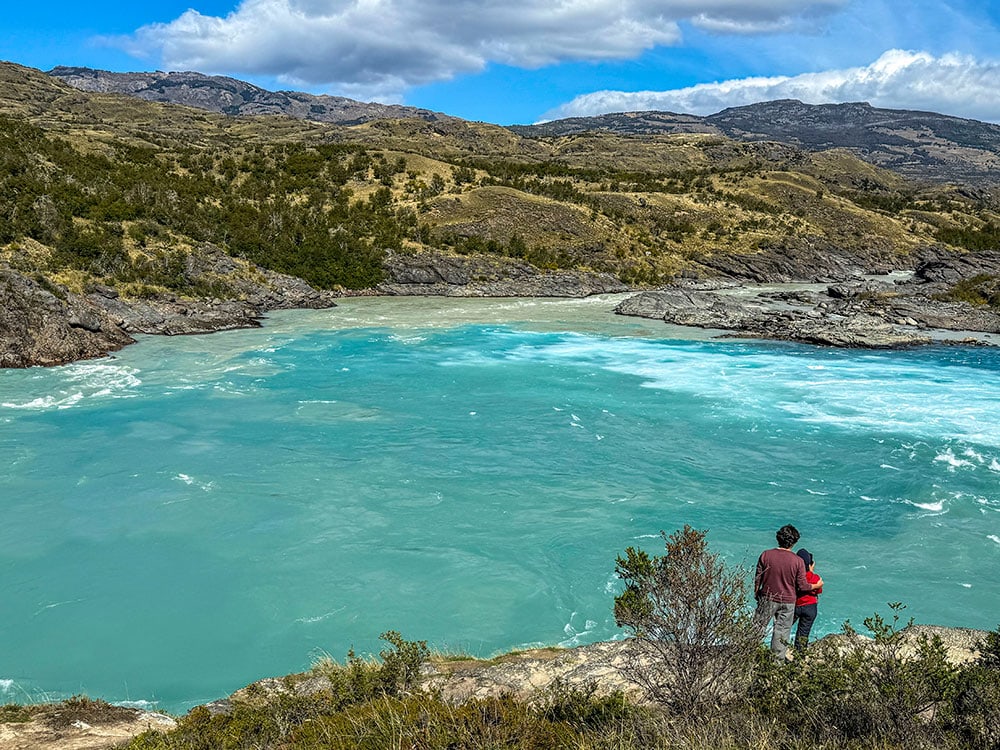 A traveler and child stand on a rocky shoreline overlooking a blue river, surrounded by hills and sparse vegetation under a partly cloudy sky, taking in the view after their hike.