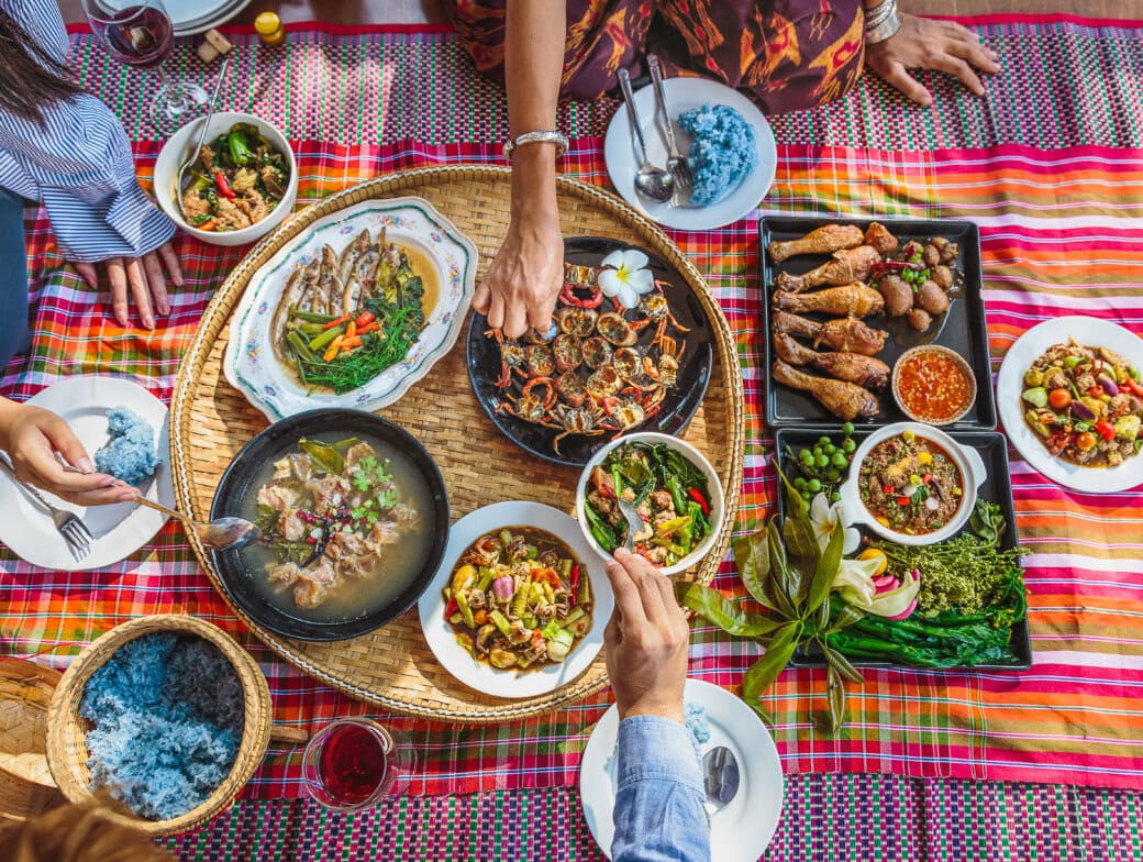 Several people sit around a woven mat, sharing a colorful spread of Thai dishes—a culinary adventure featuring rice, curries, grilled meats, vegetables, and dipping sauces.