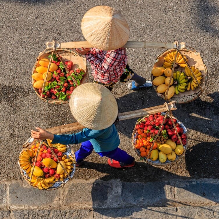 Two people wearing conical hats carry baskets filled with various fruits using shoulder poles while walking on a sunlit street, as if on a vibrant local tour.