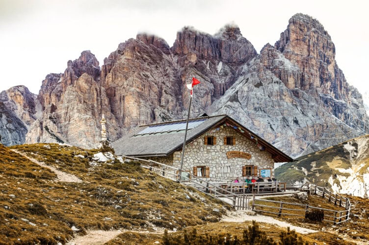 A stone mountain hut with a red flag stands in front of rugged, rocky peaks under a cloudy sky. A dirt path guides adventurers to the building, while a wooden fence surrounds the hut, inviting all on their alpine tour.