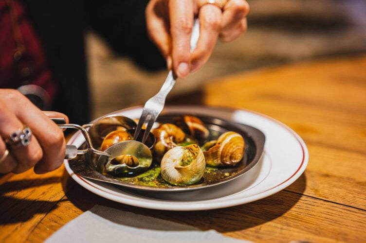 A person enjoys escargot with green herb sauce in a black dish on a wooden table, savoring this culinary adventure with special utensils.