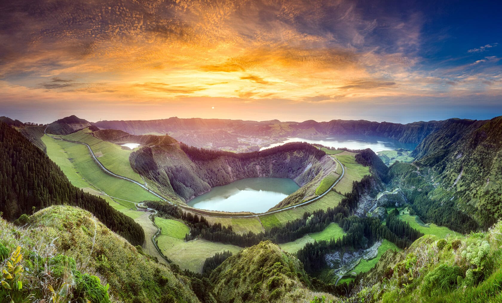 Panoramic view of a lush green volcanic crater landscape with lakes and winding roads at sunset, inviting the traveler to embrace adventure under a colorful sky.