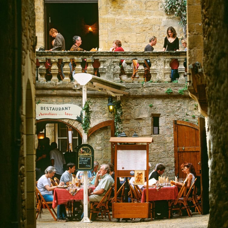 Travelers enjoy dining at tables on two levels of a rustic outdoor restaurant with stone walls and wooden accents, guided by the inviting atmosphere.