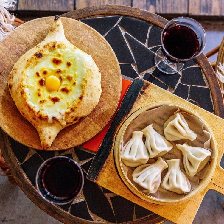 A table set for the adventurous traveler: khachapuri bread topped with egg, a plate of khinkali dumplings, and two glasses of red wine await to guide you on a delicious culinary journey.
