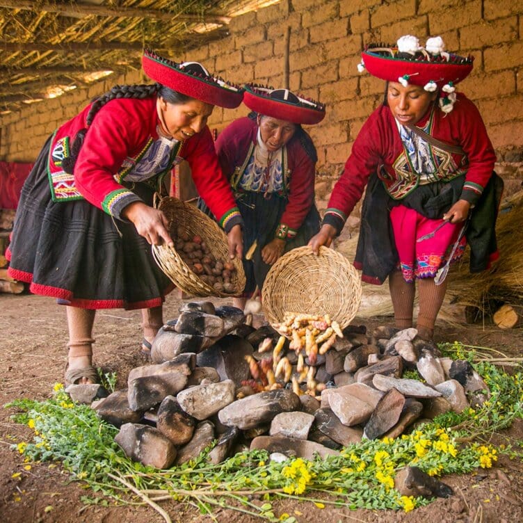 Three women in traditional Andean clothing place potatoes from baskets onto a stone oven inside a rustic building, offering travelers an authentic taste of adventure and culture.