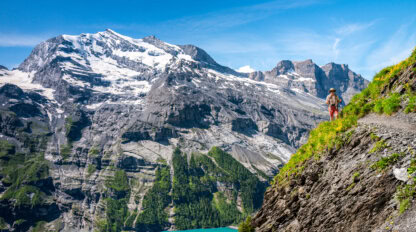 A person enjoys hiking alps on a mountain trail overlooking turquoise water, with snow-capped peaks and rocky cliffs in the background under a clear blue sky.