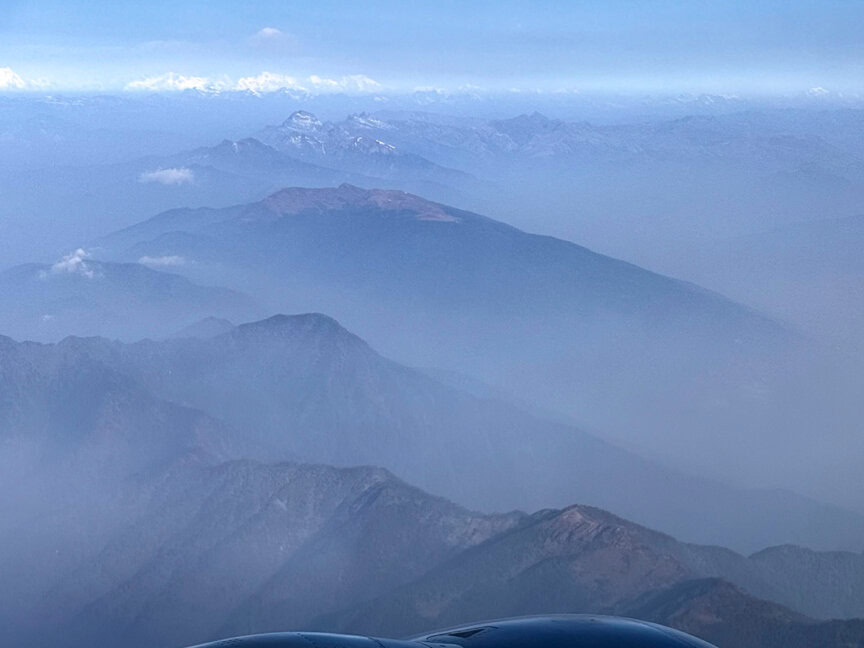 Aerial view of Bhutan’s mountain ranges with misty valleys and peaks visible in the distance under a clear blue sky, inviting a hiking adventure or cultural journey.