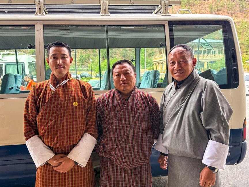 Three men wearing traditional Bhutanese attire stand in front of a white van, ready to begin their cultural journey, with buildings and greenery visible in the background.