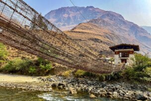 A long suspension footbridge made of rope and wood crosses a rocky river, with a traditional building and dry mountains in the background—a perfect scene for hiking in Bhutan on your cultural journey.