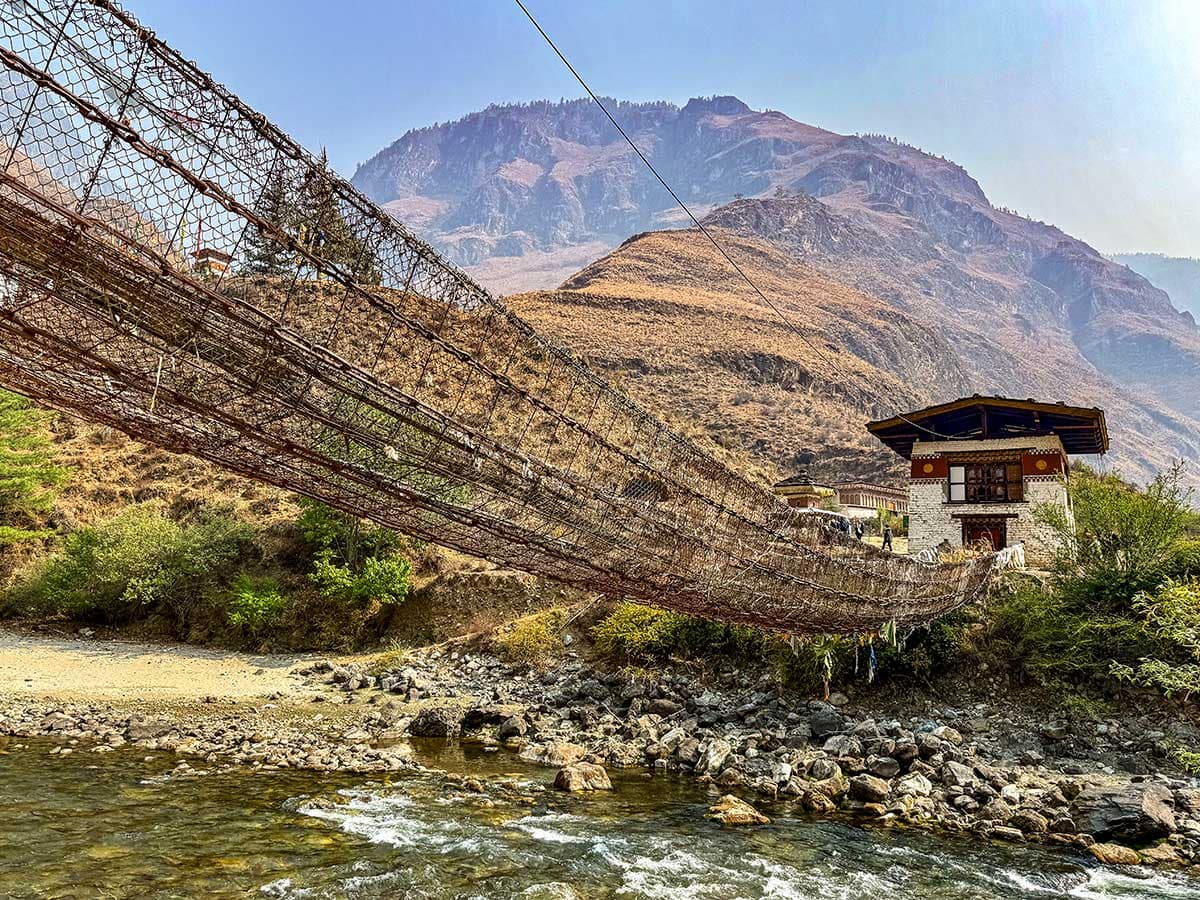 A long suspension footbridge made of rope and wood crosses a rocky river, with a traditional building and dry mountains in the background—a perfect scene for hiking in Bhutan on your cultural journey.
