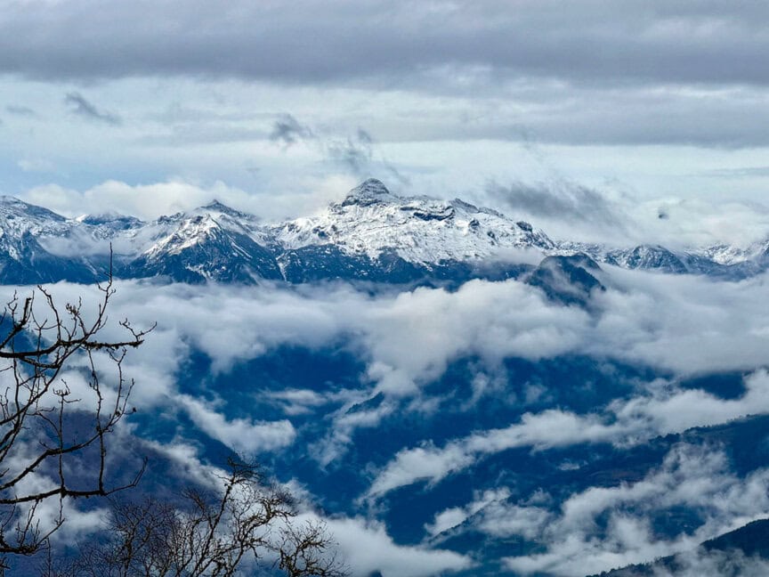 Snow-capped mountain peaks partially covered by clouds, with bare tree branches in the foreground under a cloudy sky—an iconic scene on any Hiking Bhutan adventure.