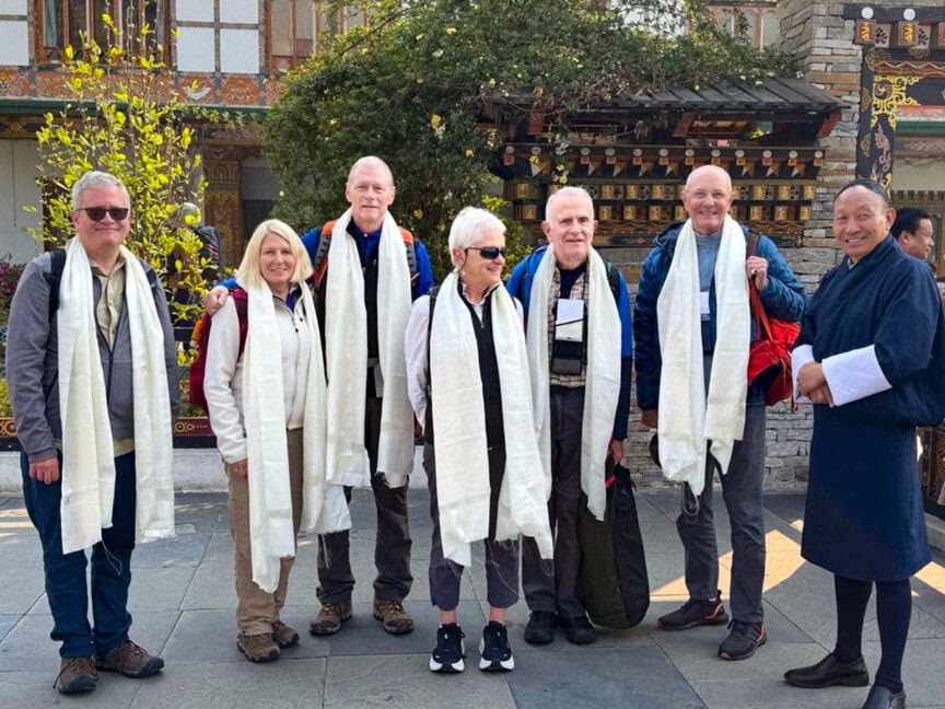 A group of six people wearing white scarves stands outdoors next to a person in traditional attire, capturing a moment from their cultural journey Bhutan, with a building and lush greenery in the background.