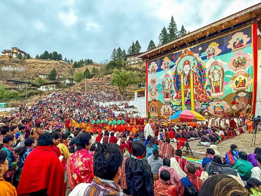 A large crowd gathers outdoors in front of a colorful, oversized Buddhist thangka for a religious festival, with monks seated and people watching the ceremony—an unforgettable highlight of any cultural journey Bhutan offers.