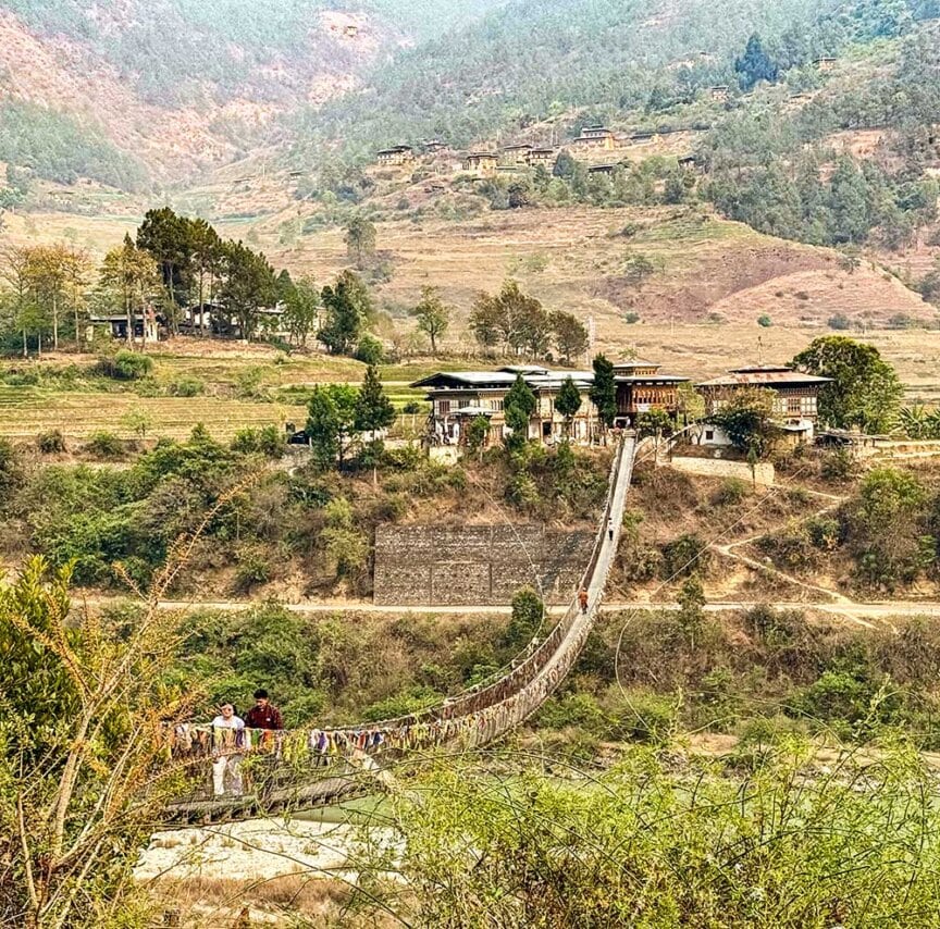 A long suspension bridge spans a valley, leading to a group of buildings, with mountains and scattered trees in the background. Several people are crossing the bridge, enjoying their Bhutan travel and the unique experience of hiking in Bhutan.