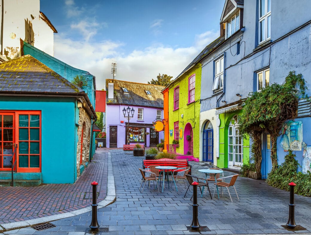 A narrow street in Cork lined with brightly painted buildings, outdoor tables and chairs, and some greenery under a partly cloudy sky—one of the top 10 Cork spots for those seeking unique things to do in Cork.