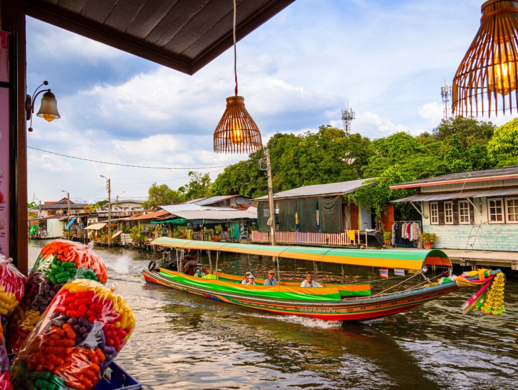 A colorful long-tail boat travels along a Bangkok canal lined with houses and greenery, viewed from a shop with snacks and ice cream signs—a perfect scene for your travel list of things to do.